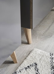 Nahaufnahme eines Möbelfußes aus Holz, der an einem grauen Möbelstück befestigt ist, auf einem hellen Holzboden mit einem Teppich im Vordergrund.