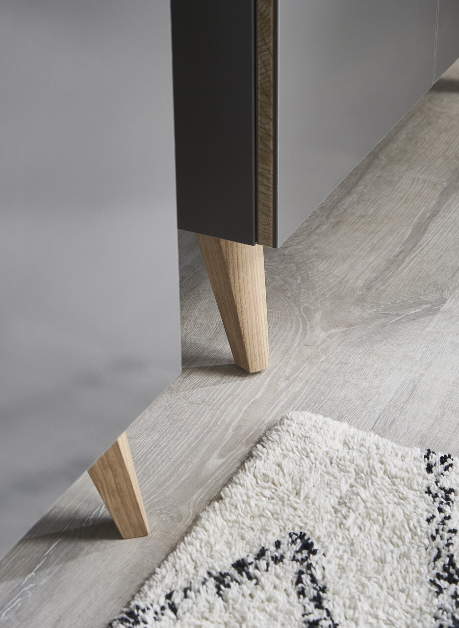 Nahaufnahme eines Möbelfußes aus Holz, der an einem grauen Möbelstück befestigt ist, auf einem hellen Holzboden mit einem Teppich im Vordergrund.