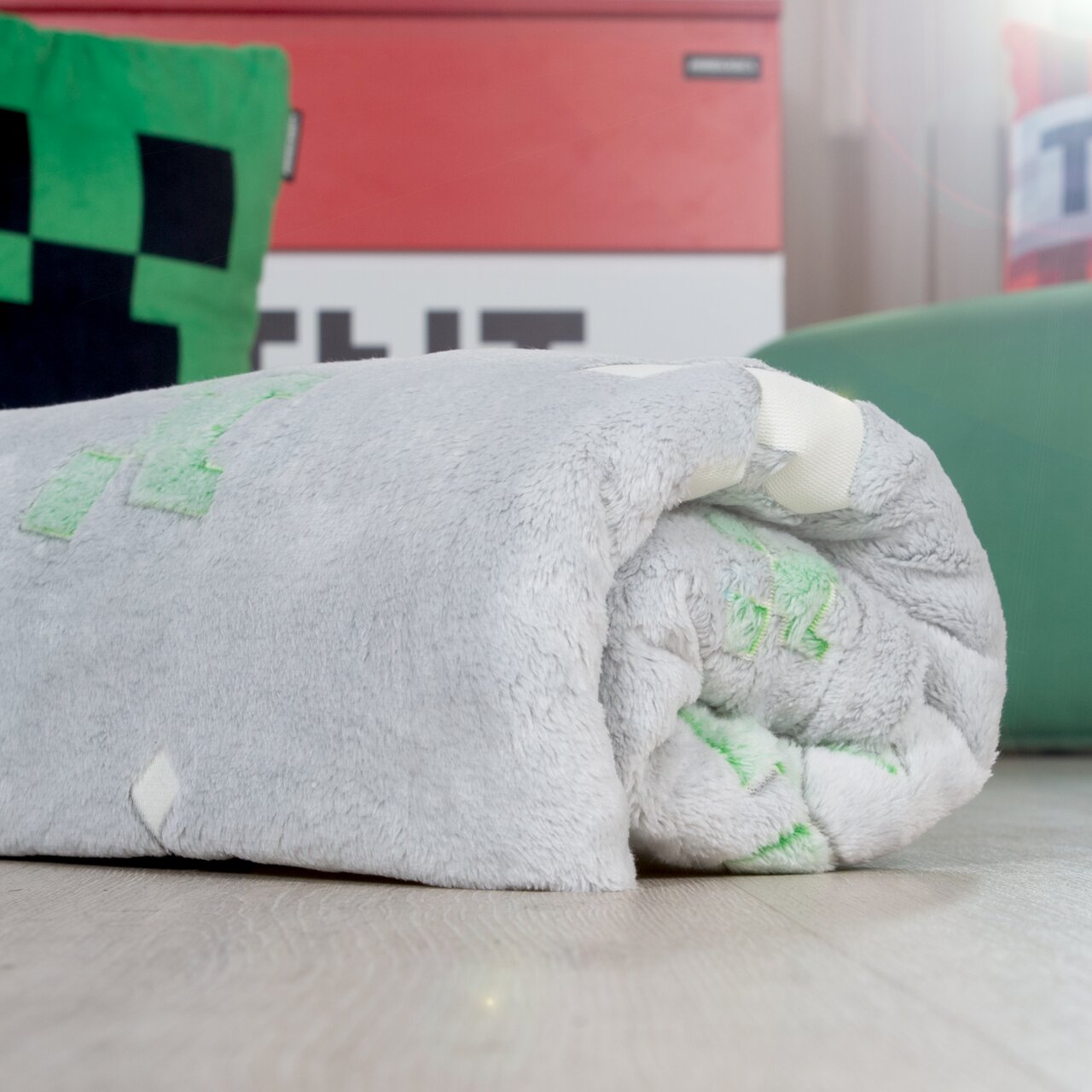 Zusammengerollte, flauschige graue Decke mit grünen Pixel-Motiven auf hellem Holzboden, seitliche Nahaufnahme aus niedriger Perspektive
