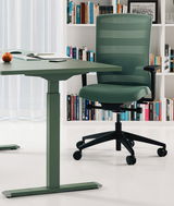 Grüner Bürostuhl mit Netzrückenlehne und schwarzen Armlehnen, seitliche Perspektive, neben einem grünen Schreibtisch in einem modernen Büro mit Bücherregal im Hintergrund.