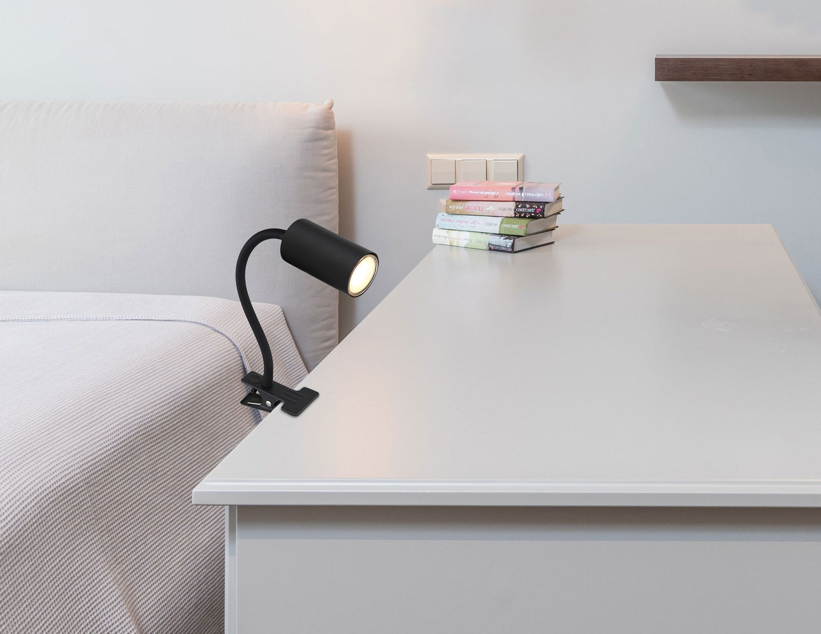 Schwarzer Klemmstrahler aus Metall, befestigt an einem Tisch neben einem Bett, seitliche Perspektive.