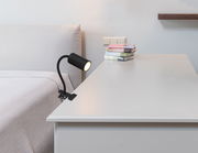 Schwarzer Klemmstrahler aus Metall, befestigt an einem Tisch neben einem Bett, seitliche Perspektive.