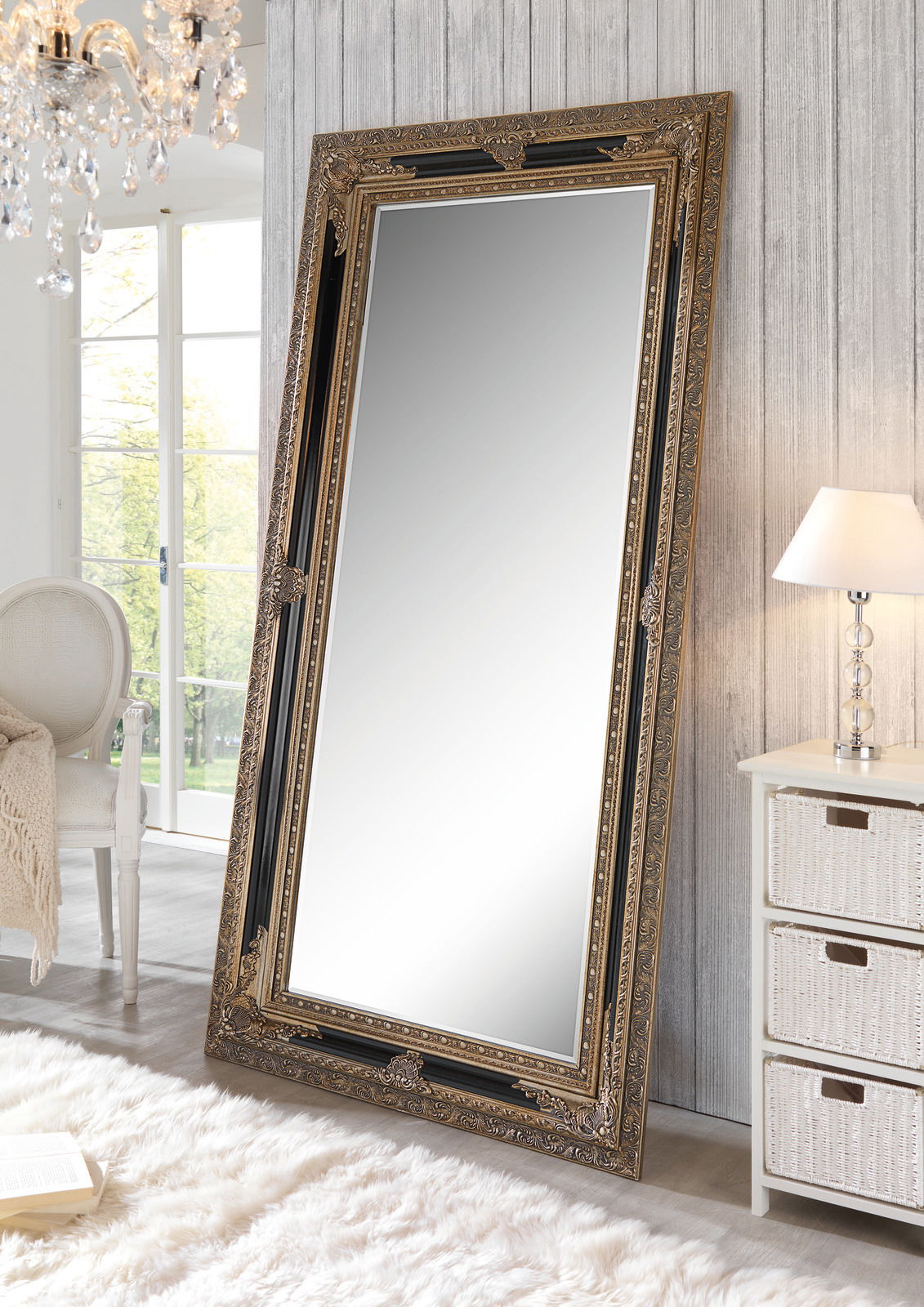 Großer, reich verzierter Spiegel im klassischen Stil, der an eine Wand gelehnt ist. Der Spiegel hat einen goldenen Rahmen und steht in einem hellen, elegant eingerichteten Raum.