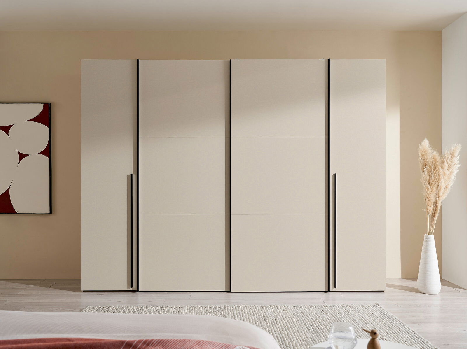 Moderner, breiter Kleiderschrank in hellem Beige mit vier glatten Fronten und langen schwarzen Griffleisten, frontal aus Augenhöhe im Schlafzimmer fotografiert.