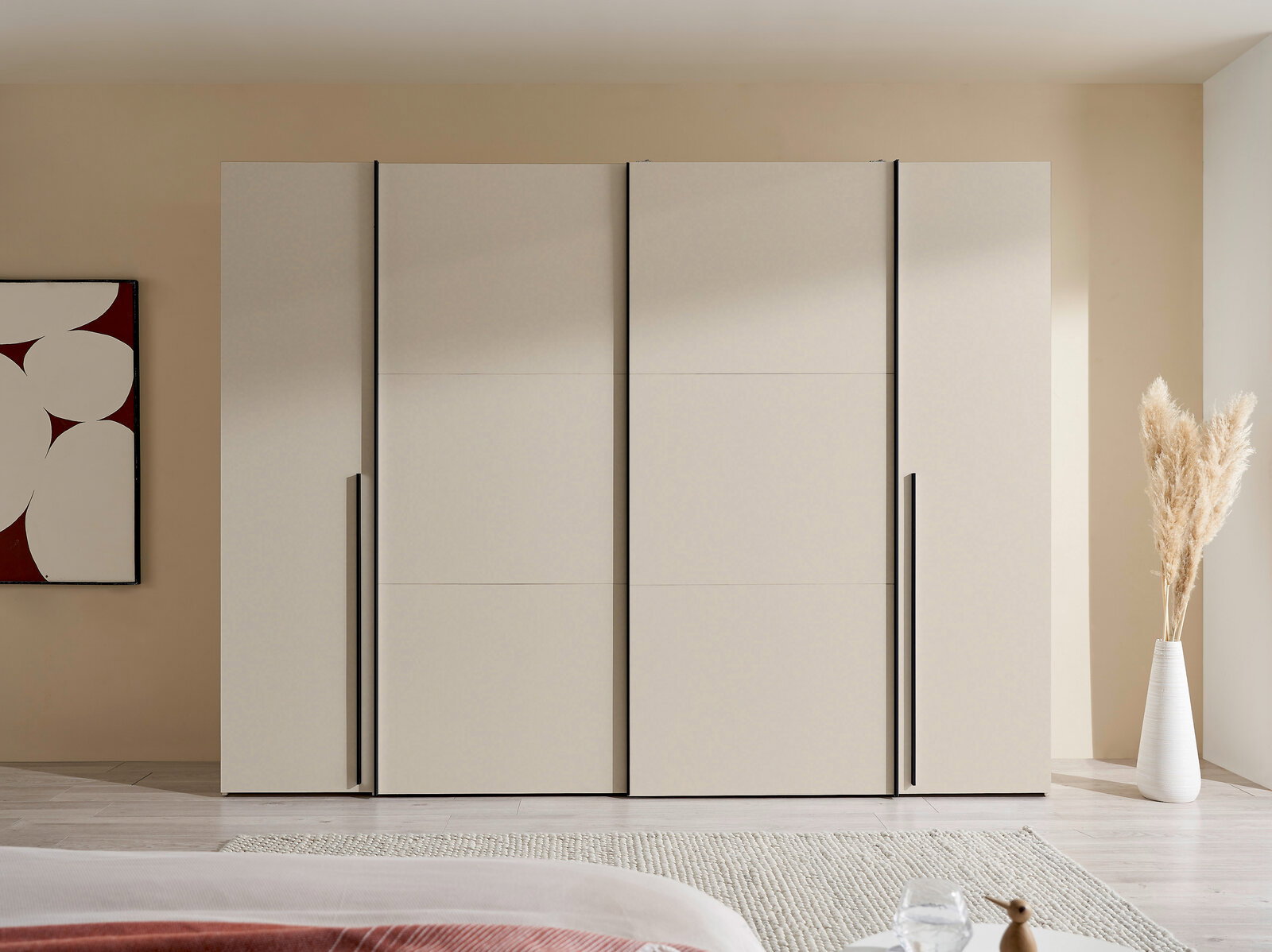 Moderner, breiter Kleiderschrank in hellem Beige mit vier glatten Fronten und langen schwarzen Griffleisten, frontal aus Augenhöhe im Schlafzimmer fotografiert.