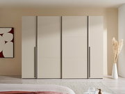 Moderner, breiter Kleiderschrank in hellem Beige mit vier glatten Fronten und langen schwarzen Griffleisten, frontal aus Augenhöhe im Schlafzimmer fotografiert.