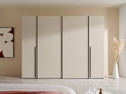 Moderner, breiter Kleiderschrank in hellem Beige mit vier glatten Fronten und langen schwarzen Griffleisten, frontal aus Augenhöhe im Schlafzimmer fotografiert.