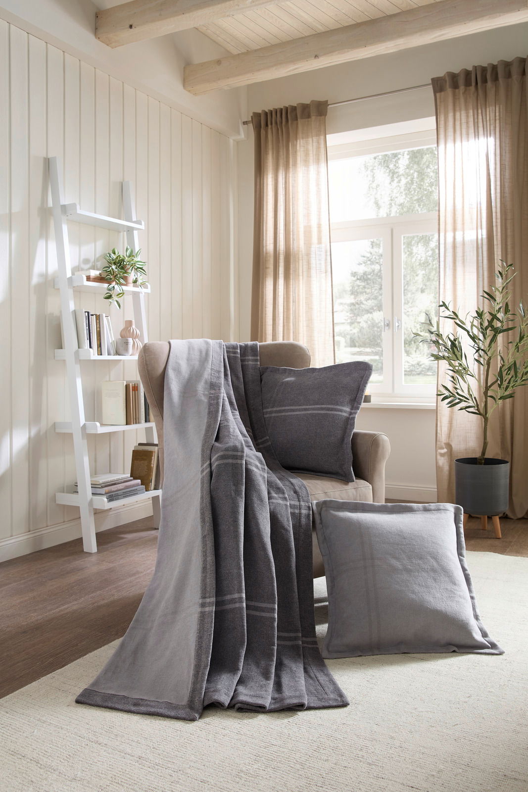Linneweber Wohndecke BRAGA Gemütliches Wohnzimmer mit grauer Wohndecke und Kissen auf einem Sessel, im Hintergrund ein Regal und ein Fenster mit Vorhängen, seitliche Perspektive.