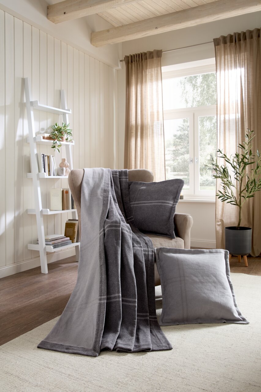 Linneweber Wohndecke BRAGA Gemütliches Wohnzimmer mit grauer Wohndecke und Kissen auf einem Sessel, im Hintergrund ein Regal und ein Fenster mit Vorhängen, seitliche Perspektive.