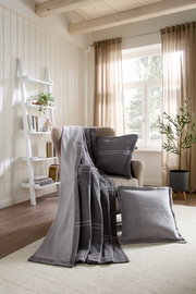 Gemütliches Wohnzimmer mit grauer Wohndecke und Kissen auf einem Sessel, im Hintergrund ein Regal und ein Fenster mit Vorhängen, seitliche Perspektive.
