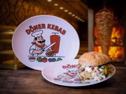 Weißer Döner-Kebab-Teller mit Cartoon-Koch-Logo und Schriftzug „Döner Kebab“, vorne ein gefüllter Döner-Burger auf dem Teller; Perspektive: leicht von vorne in Augenhöhe, mit unscharfem Grillspieß im Hintergrund.