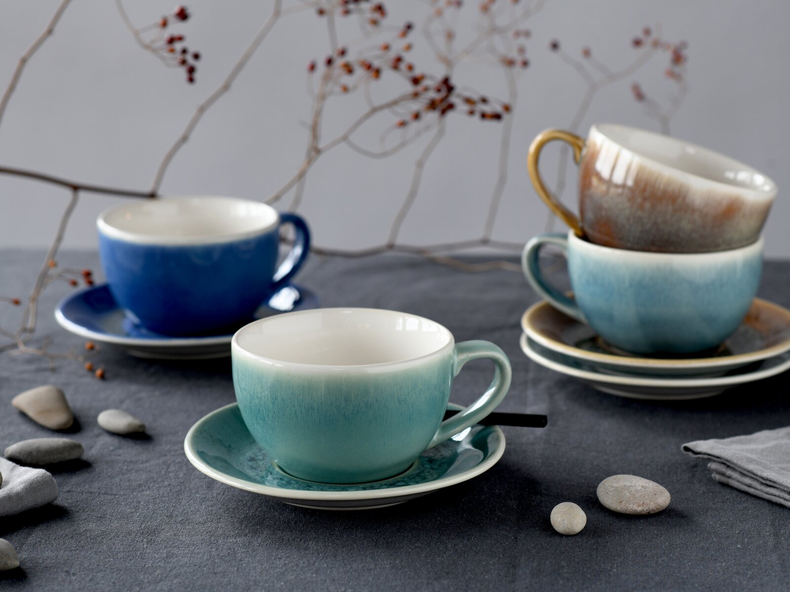 Stilvolles Keramik-Tassen- und Untertassen-Set in Blau- und Türkistönen, eine Tasse mit goldfarbenem Henkel, arrangiert auf grauem Tisch mit Dekoästen – Perspektive: leicht schräg von vorne auf Tischhöhe