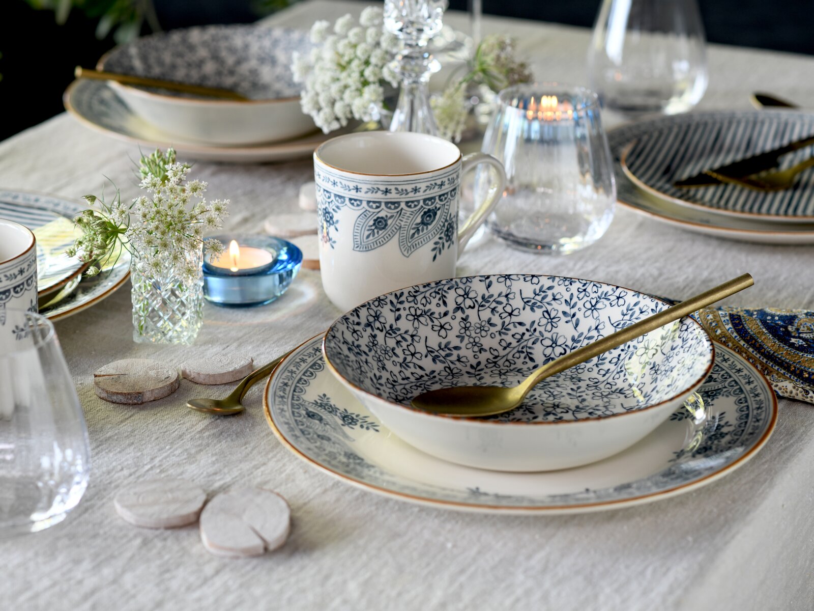 Schick gedeckter Esstisch mit weiß-blau gemusterter Keramikschale auf passendem Teller, goldfarbenem Löffel und Tasse, umgeben von Gläsern, Kerzenlicht und Blumen, aus leicht seitlicher Nahaufnahme