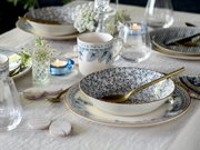 Schick gedeckter Esstisch mit weiß-blau gemusterter Keramikschale auf passendem Teller, goldfarbenem Löffel und Tasse, umgeben von Gläsern, Kerzenlicht und Blumen, aus leicht seitlicher Nahaufnahme