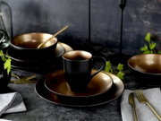 Schwarzes Keramik-Geschirrset mit goldfarbenem Innenfinish: Tasse auf Teller, daneben Schalen und Besteck, fotografiert in leicht seitlicher Perspektive auf einem dunklen Tisch.