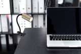 Klemmstrahler aus Metall in altmessing Optik, seitliche Perspektive, befestigt an einem Tisch neben einem Laptop.