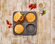 Vogelperspektive auf ein Burger Brötchen-Blech mit drei runden Formen, zwei mit gebackenen Brötchen und eine leer, umgeben von frischen Zutaten wie Tomaten, Zwiebeln und Sesam.