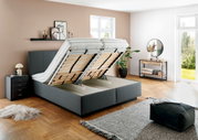 Graues Polsterbett mit hochgeklappten Lattenrosten, seitliche Perspektive, modernes Schlafzimmerdesign mit Nachttisch und Dekoration.