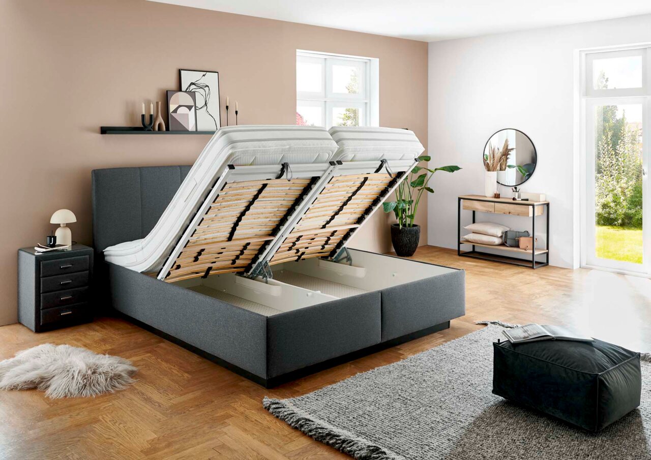 Graues Polsterbett mit hochgeklappten Lattenrosten, seitliche Perspektive, modernes Schlafzimmerdesign mit Nachttisch und Dekoration.