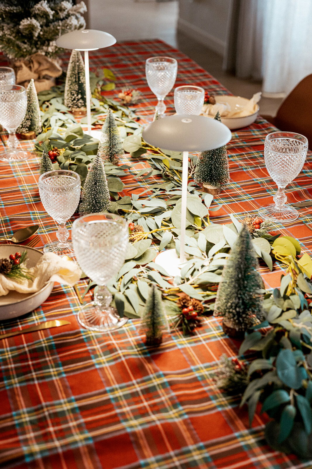 Festlich gedeckter Esstisch mit rot kariertem Tischtuch, Eukalyptus-Girlande, kleinen Deko-Tannenbäumen, Kristallgläsern und zwei weißen Tischlampen, fotografiert aus schräger Vogelperspektive.