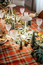 Festlich gedeckter Esstisch mit rot kariertem Tischtuch, Eukalyptus-Girlande, kleinen Deko-Tannenbäumen, Kristallgläsern und zwei weißen Tischlampen, fotografiert aus schräger Vogelperspektive.