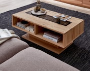 Moderner Couchtisch aus hellem Massivholz mit dunkler Glas- oder Stein-Einlage und offenen Ablagefächern, dekoriert mit Tablett, Gläsern und Büchern, in schräger Draufsicht im Wohnzimmer auf Teppich.