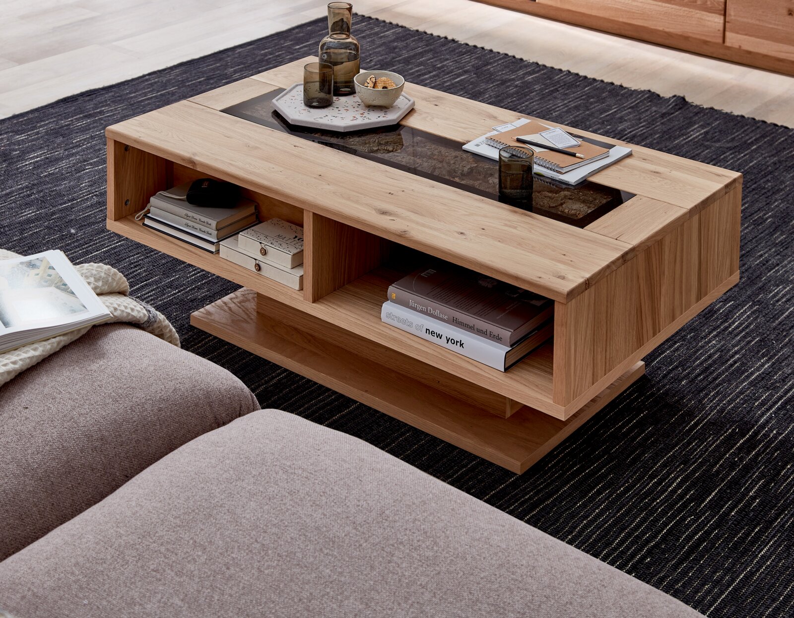 Cantus Couchtisch DONNA Moderner Couchtisch aus hellem Massivholz mit dunkler Glas- oder Stein-Einlage und offenen Ablagefächern, dekoriert mit Tablett, Gläsern und Büchern, in schräger Draufsicht im Wohnzimmer auf Teppich.