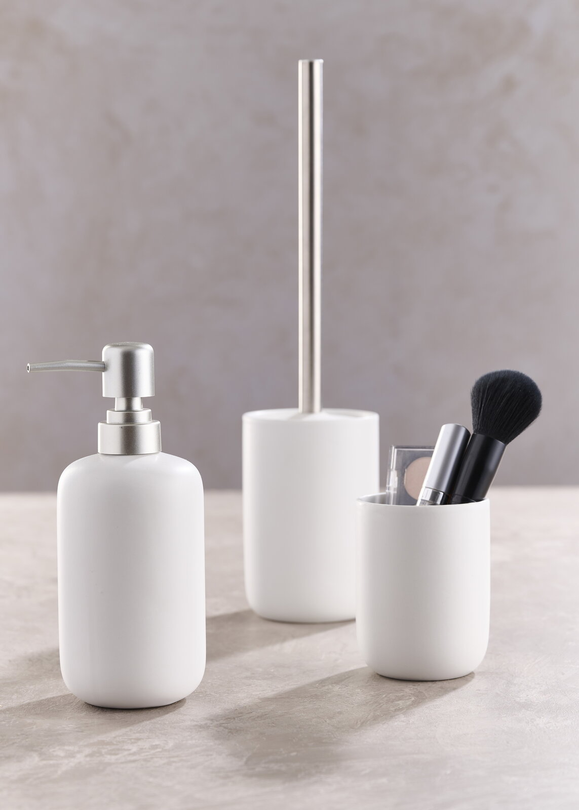 Dreiteiliges Badset in Weiß, bestehend aus einem Seifenspender, einer Toilettenbürste und einem Becher mit Make-up-Pinsel, aus seitlicher Perspektive fotografiert.