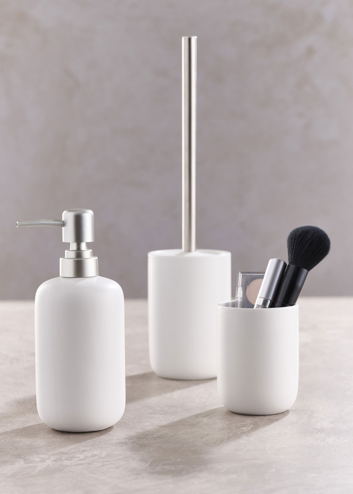 Dreiteiliges Badset in Weiß, bestehend aus einem Seifenspender, einer Toilettenbürste und einem Becher mit Make-up-Pinsel, aus seitlicher Perspektive fotografiert.
