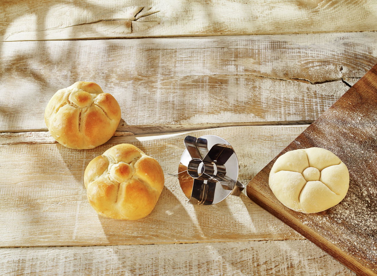 Brötchenstempel aus Metall auf einem Holztisch, umgeben von geformten und ungeformten Brötchen. Draufsicht.