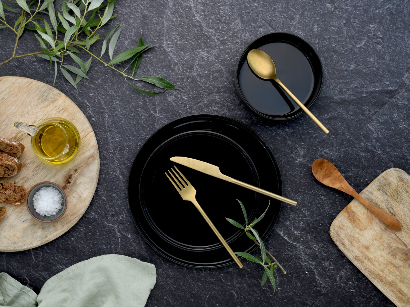 Schwarzes Geschirr-Set mit goldfarbenem Besteck (Teller und Schale) auf dunkler Steinoberfläche, dekoriert mit Olivenzweigen, Brot, Salz und Öl – Draufsicht (Flatlay).