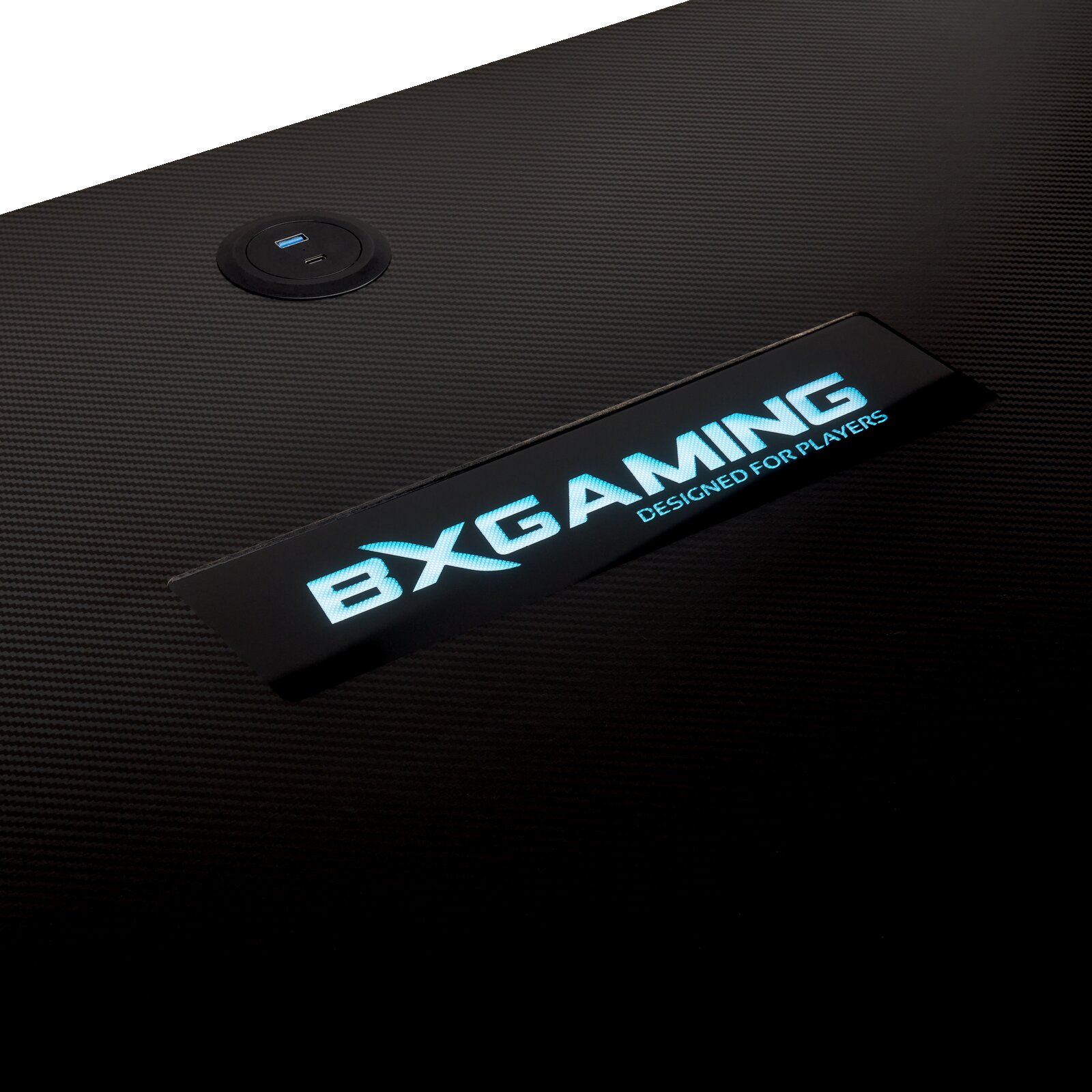 Nahaufnahme einer schwarzen, carbonähnlich strukturierten Gaming-Tischoberfläche mit integriertem runden USB-Anschluss und leuchtendem BXGAMING-Schriftzug, aufgenommen aus schräger Draufsicht.