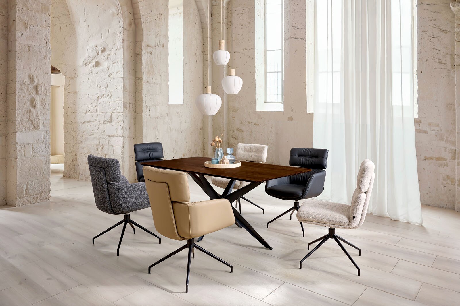Dieter Knoll Eettafel TAVOLO Moderne eethoek met donkere houten eettafel en zes gestoffeerde draaistoelen in verschillende kleuren, gefotografeerd schuin van voren op ooghoogte in een lichte loft met witte bakstenen muren en grote ramen.