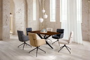 Modernes Esszimmer-Set mit rechteckigem Esstisch in dunkler Holzoptik und sechs gepolsterten Drehstühlen in verschiedenen Farben, fotografiert in schräger Vorderansicht in einem hellen Loft mit hohen Fenstern und weißen Steinwänden