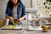 Weiße Küchenmaschine in Seitenansicht mit Rührschüssel und Knethaken, im Hintergrund eine Person beim Backen.