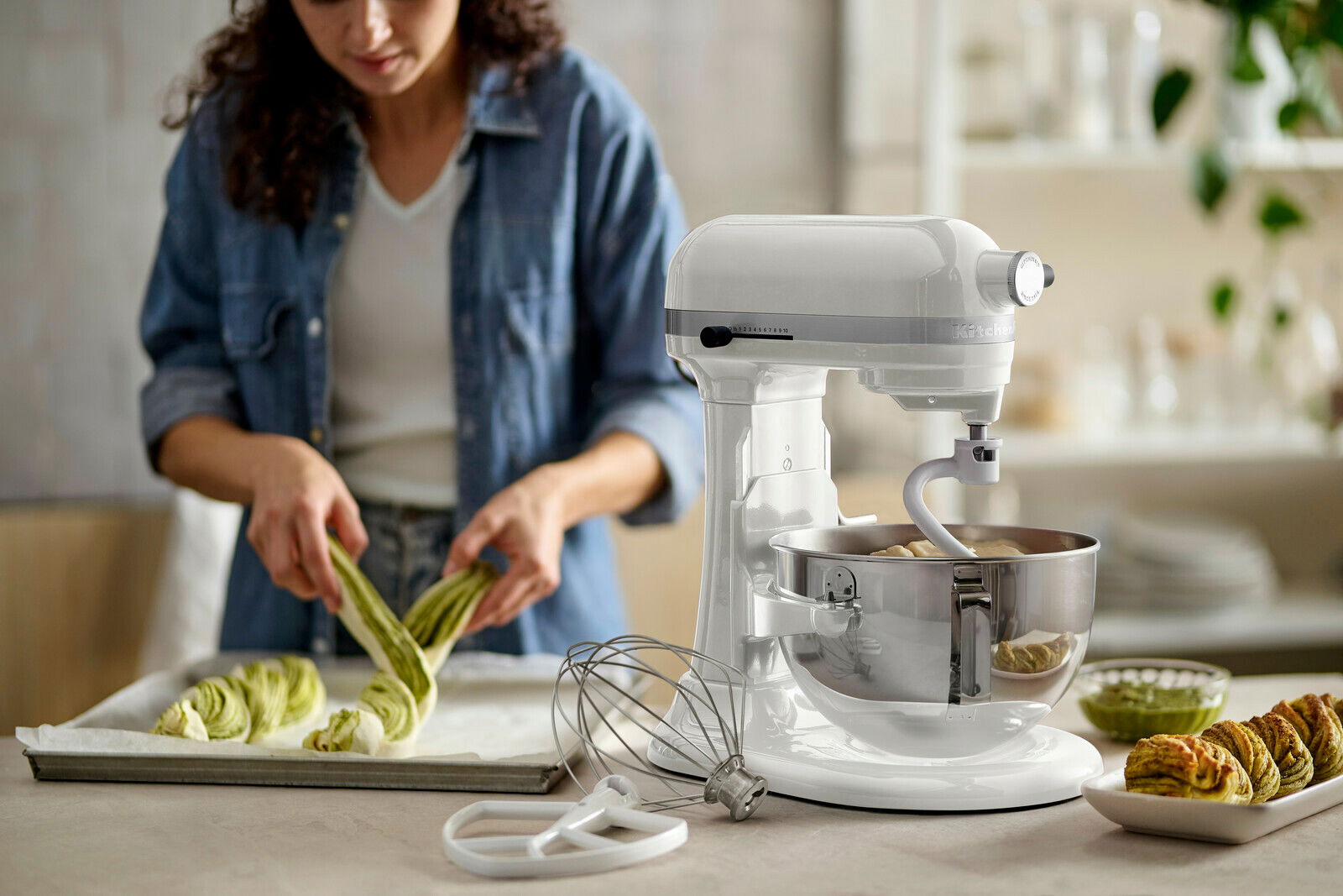 Weiße Küchenmaschine in Seitenansicht mit Rührschüssel und Knethaken, im Hintergrund eine Person beim Backen.
