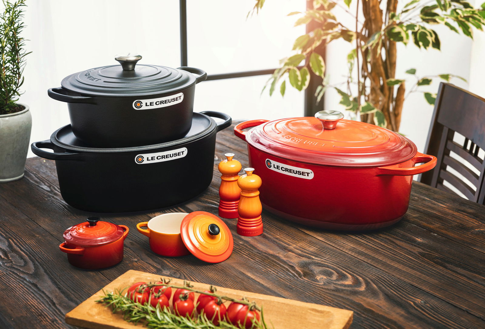 Verschiedene Le Creuset Kochgeschirre in Kirschrot und Schwarz auf einem Holztisch, fotografiert aus einer leicht erhöhten Perspektive. Im Vordergrund eine Mini-Cocotte, im Hintergrund ein großer ovaler Bräter und ein runder Bräter, umgeben von Gewürzmühl