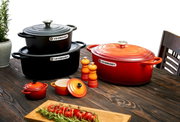 Verschiedene Le Creuset Kochgeschirre in Kirschrot und Schwarz auf einem Holztisch, fotografiert aus einer leicht erhöhten Perspektive. Im Vordergrund eine Mini-Cocotte, im Hintergrund ein großer ovaler Bräter und ein runder Bräter, umgeben von Gewürzmühl