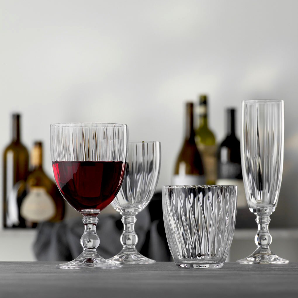 Vier elegante Gläser aus klarem Glas, darunter ein Weinkelch mit rotem Wein, ein kleinerer Weinkelch, ein Becher und ein Sektkelch, auf einem Tisch mit unscharfem Hintergrund von Weinflaschen. Frontale Perspektive.