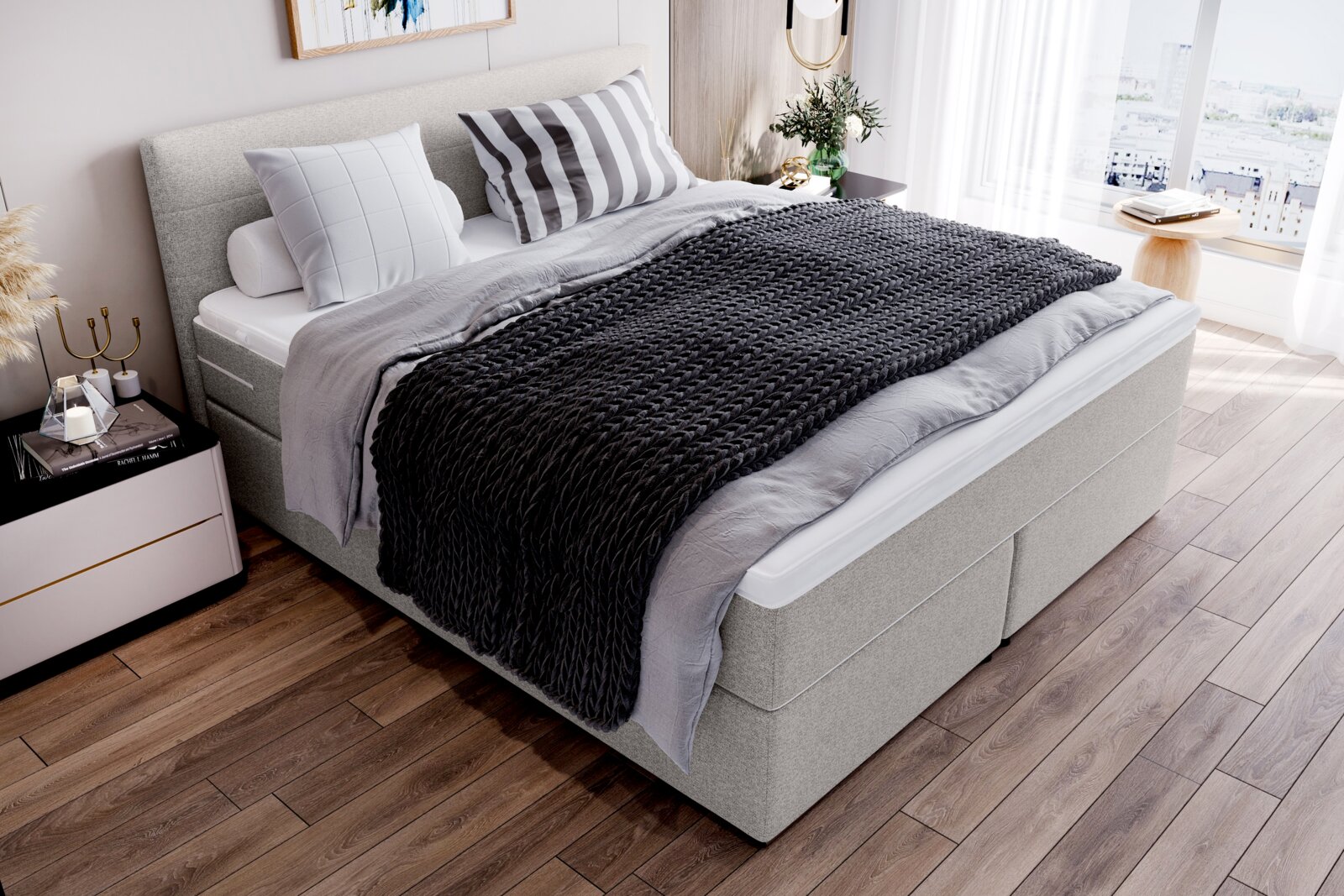 Gepolstertes Boxspringbett in hellgrauem Stoff mit hohem Kopfteil, weißer Matratzenauflage und dunkelgrauer grob gestrickter Tagesdecke im hellen Schlafzimmer, Perspektive: schräg von vorne links (leicht von oben)