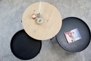 Draufsicht auf einen runden Couchtisch mit einer hellen Holzplatte und zwei schwarzen Platten, dekoriert mit Kerzen und einer Zeitschrift.