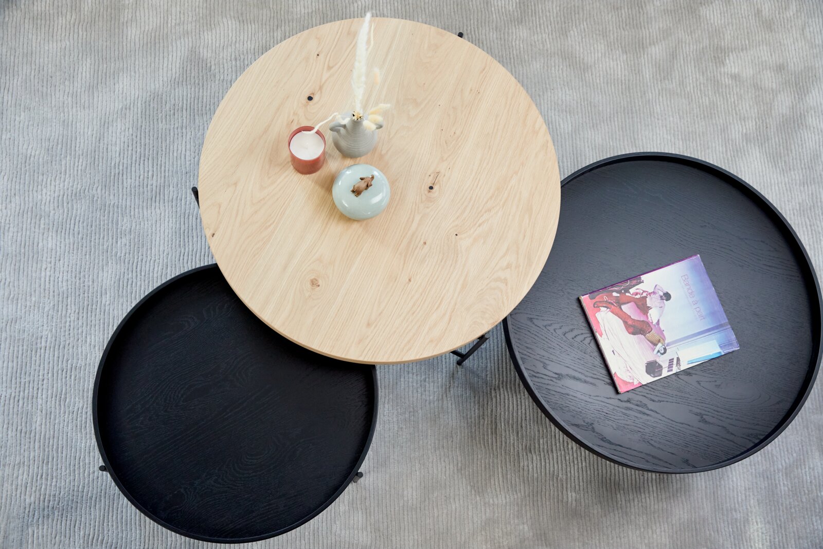 Draufsicht auf einen runden Couchtisch mit einer hellen Holzplatte und zwei schwarzen Platten, dekoriert mit Kerzen und einer Zeitschrift.