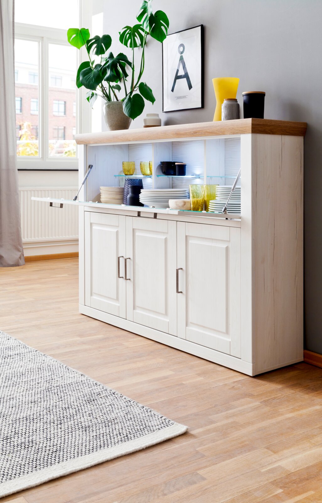Weißes Sideboard im Landhausstil mit naturfarbener Holzplatte und Klapptür mit Glasböden, teils geöffnet und mit Geschirr dekoriert, in heller Wohnzimmerecke – Perspektive: leicht von vorne rechts.