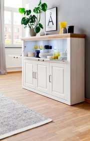 Weißes Sideboard im Landhausstil mit naturfarbener Holzplatte und Klapptür mit Glasböden, teils geöffnet und mit Geschirr dekoriert, in heller Wohnzimmerecke – Perspektive: leicht von vorne rechts.