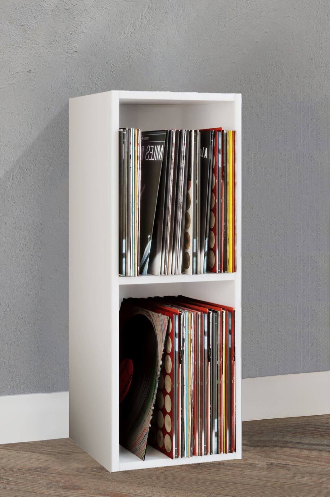 Weißes, schmales Vinyl-Schallplattenregal mit zwei Fächern, gefüllt mit LPs, auf Holzboden vor grauer Wand, in leicht schräger Frontansicht (3/4-Perspektive) von vorn links.