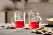 Zwei transparente Glasbecher mit Henkeln und dampfendem rotem Tee, frontal in stimmungsvoller Tischszene betrachtet
