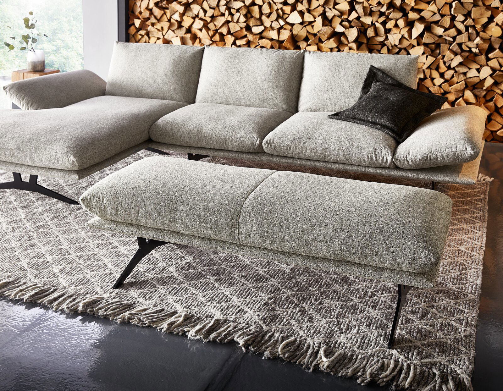 Hellgraues, modernes Ecksofa mit Chaiselongue links und passendem Hocker auf gemustertem Teppich, seitliche Perspektive leicht von vorne; im Hintergrund gestapeltes Kaminholz und ein schwarzes Kissen.