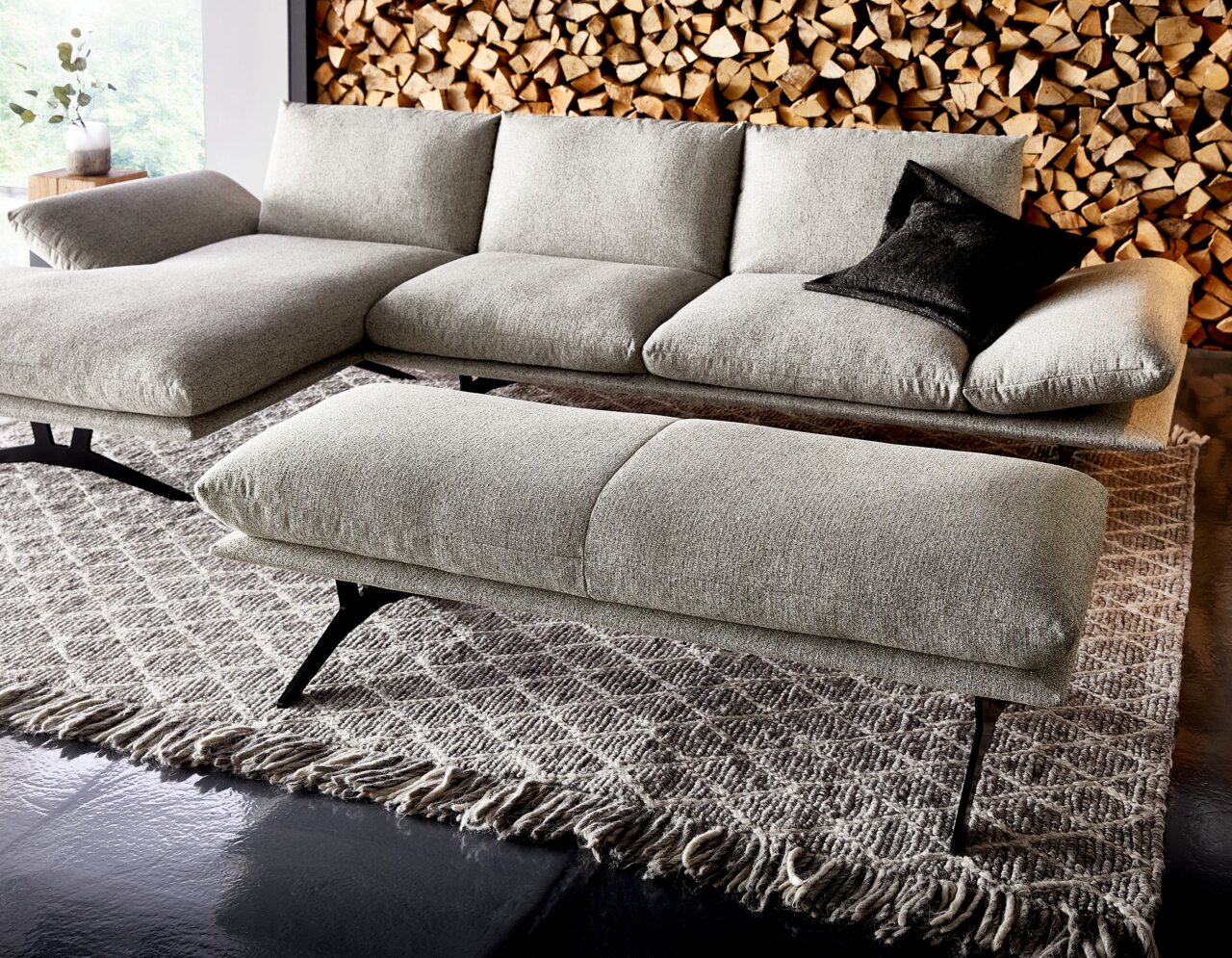 Hellgraues, modernes Ecksofa mit Chaiselongue links und passendem Hocker auf gemustertem Teppich, seitliche Perspektive leicht von vorne im Hintergrund gestapeltes Kaminholz und ein schwarzes Kissen.