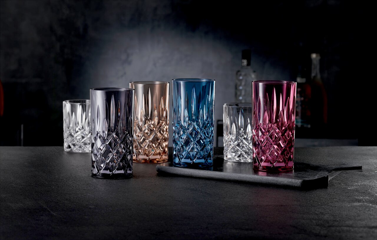 Veelkleurige kristallen drinkglazen met facetten (blauw, roze, rookgrijs, amber en helder) op een donker barblad, van voren gefotografeerd vanuit een licht verhoogd perspectief.