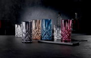 Veelkleurige kristallen drinkglazen met facetten (blauw, roze, rookgrijs, amber en helder) op een donker barblad, van voren gefotografeerd vanuit een licht verhoogd perspectief.